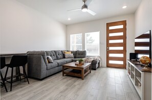 A modern living room with a grey sectional sofa, a wooden coffee table, a black bar stool, a wooden door, and a television on a stand. - Brand New Home Near Downtown (Sulphur Springs)