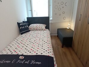 Ein Bett mit einem Punkte-Bettbezug, ein Kissen mit dem Aufdruck 'Welcome & Feel at Home', ein Nachttisch mit einer Lampe und eine Wand mit sechseckigen Mustern.