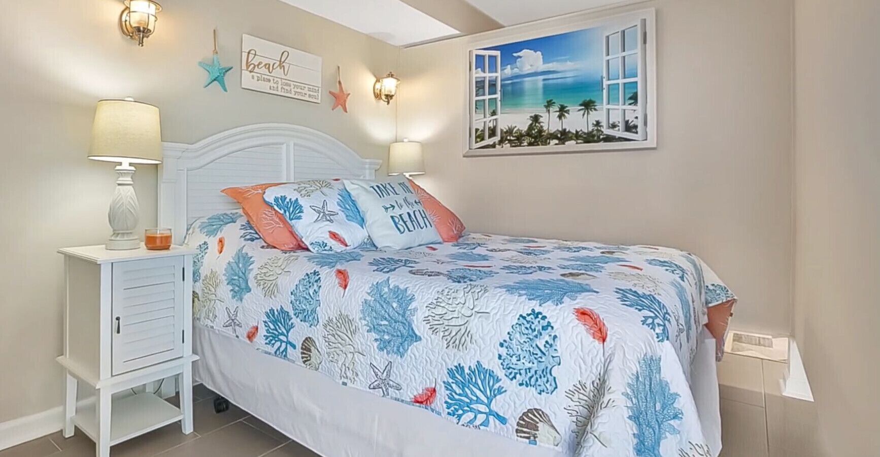 Un dormitorio con cama, mesitas de noche, una lámpara de pared, una decoración de estrella de mar, un cuadro con motivos playeros y una colcha con tema de playa.