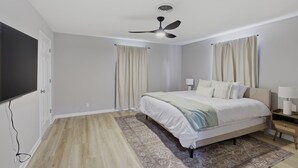 Un dormitorio con cama, ventilador de techo, mesita de noche con lámpara y televisor montado en la pared.