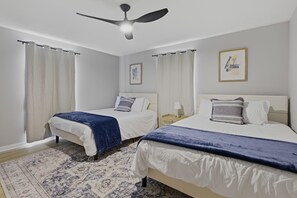 Une chambre avec deux lits, un ventilateur de plafond, des rideaux, un tapis et deux tableaux encadrés au mur.