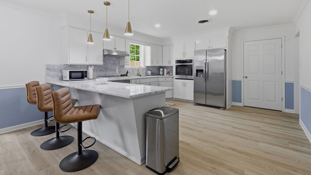 Una cocina moderna con una isla central, taburetes de bar, electrodomésticos de acero inoxidable, iluminación colgante y vistas al exterior.
