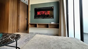 A modern living room with a large flat-screen TV mounted on a green wall, wooden shelving below, and a large window to the right.