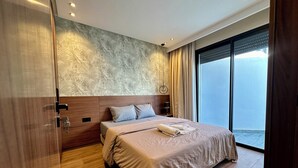 A modern bedroom with a large bed, a headboard, a nightstand, a wall-mounted lamp, and a view of the outside.