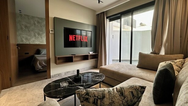A modern living room with a large flat-screen TV, a sofa, a coffee table, and a sliding glass door leading to a balcony.