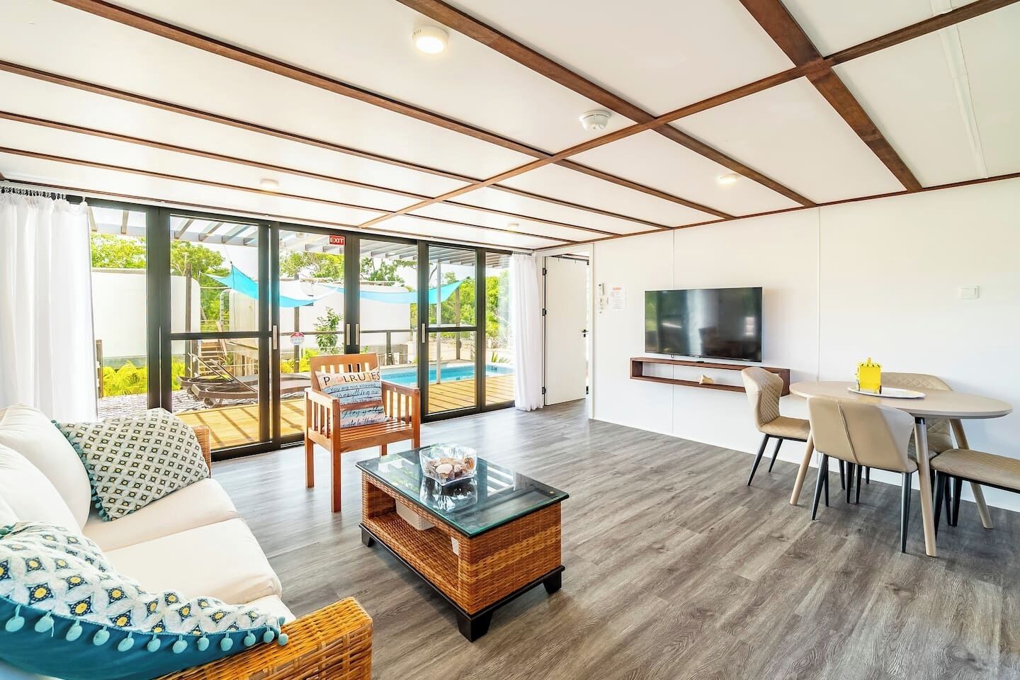 A spacious living area with a sofa, coffee table, and dining set. A TV is mounted on the wall, and a pool is visible through large windows.