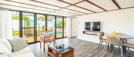Amplia sala de estar con sofá, mesa de centro y comedor. Un televisor está montado en la pared, y se ve una piscina a través de grandes ventanales.
