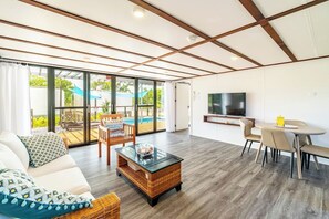 A spacious living area with a sofa, coffee table, and dining set. A TV is mounted on the wall, and a pool is visible through large windows.