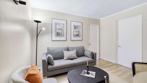 Ett modernt vardagsrum med en grå soffa, ett svart soffbord och en golvlampa. På väggen hänger två inramade tavlor, och i bakgrunden syns en dörr.