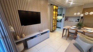 Un salon moderne doté d’un téléviseur à écran plat, d’un coin repas avec une table dressée pour deux personnes, et d’une cuisine en arrière-plan.