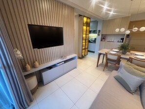 Ein modernes Wohnzimmer mit Flachbildfernseher, einem Essbereich mit Tisch für zwei Personen und einer Küche im Hintergrund.