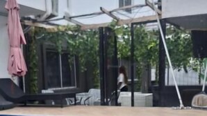 Una piscina con solárium de baldosas, un área de estar cubierta y un edificio blanco al fondo.