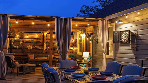 Un espace extérieur aménagé pour les repas, avec une table dressée pour quatre personnes, entourée de chaises, et un coin salon abrité avec des coussins et une lampe.