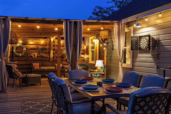 An outdoor dining area with a table set for four, surrounded by chairs, and a covered seating area with cushions and a lamp. - Lovely 2-bedroom waterfront cottage with many outdoor activities and charm (Foley)