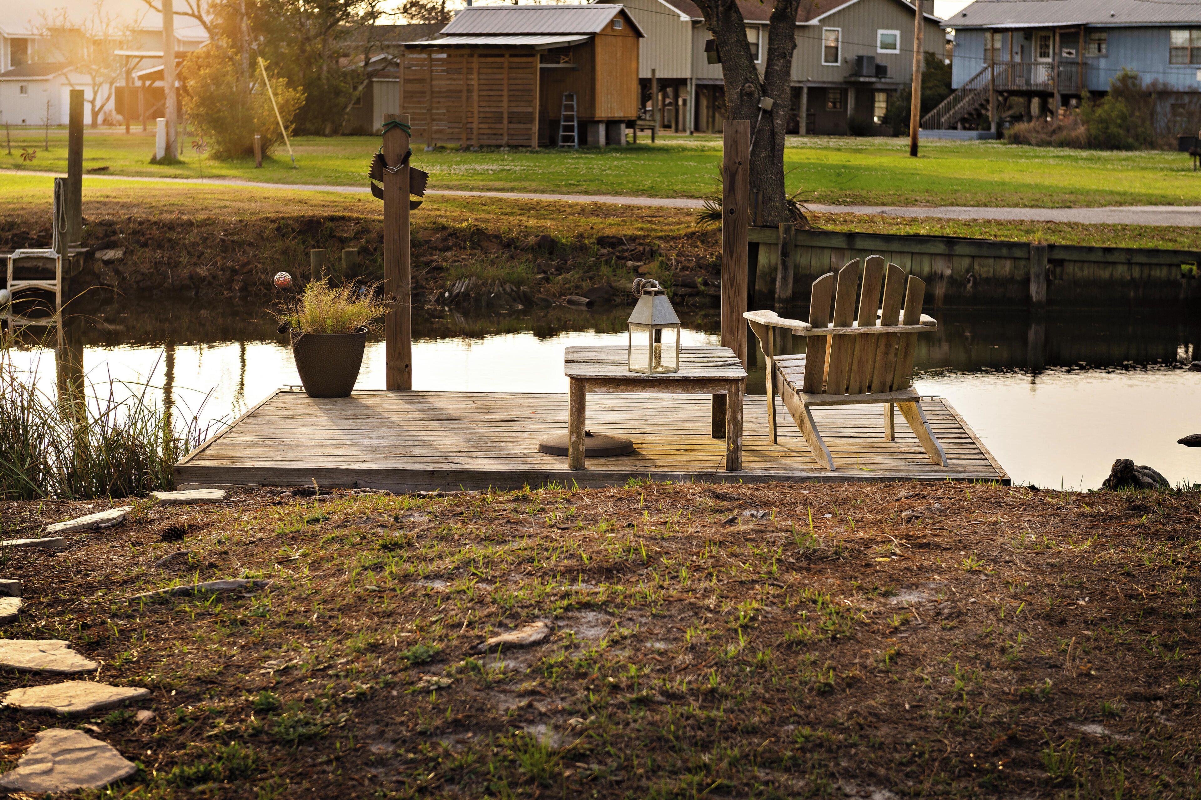 A wooden dock with a table and chairs, a lantern, and a potted plant by a calm water body.