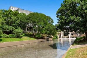 A calm river with a concrete walkway alongside, surrounded by lush greenery and a building in the background. - BLOCK La Cascada 2br (Orlando)