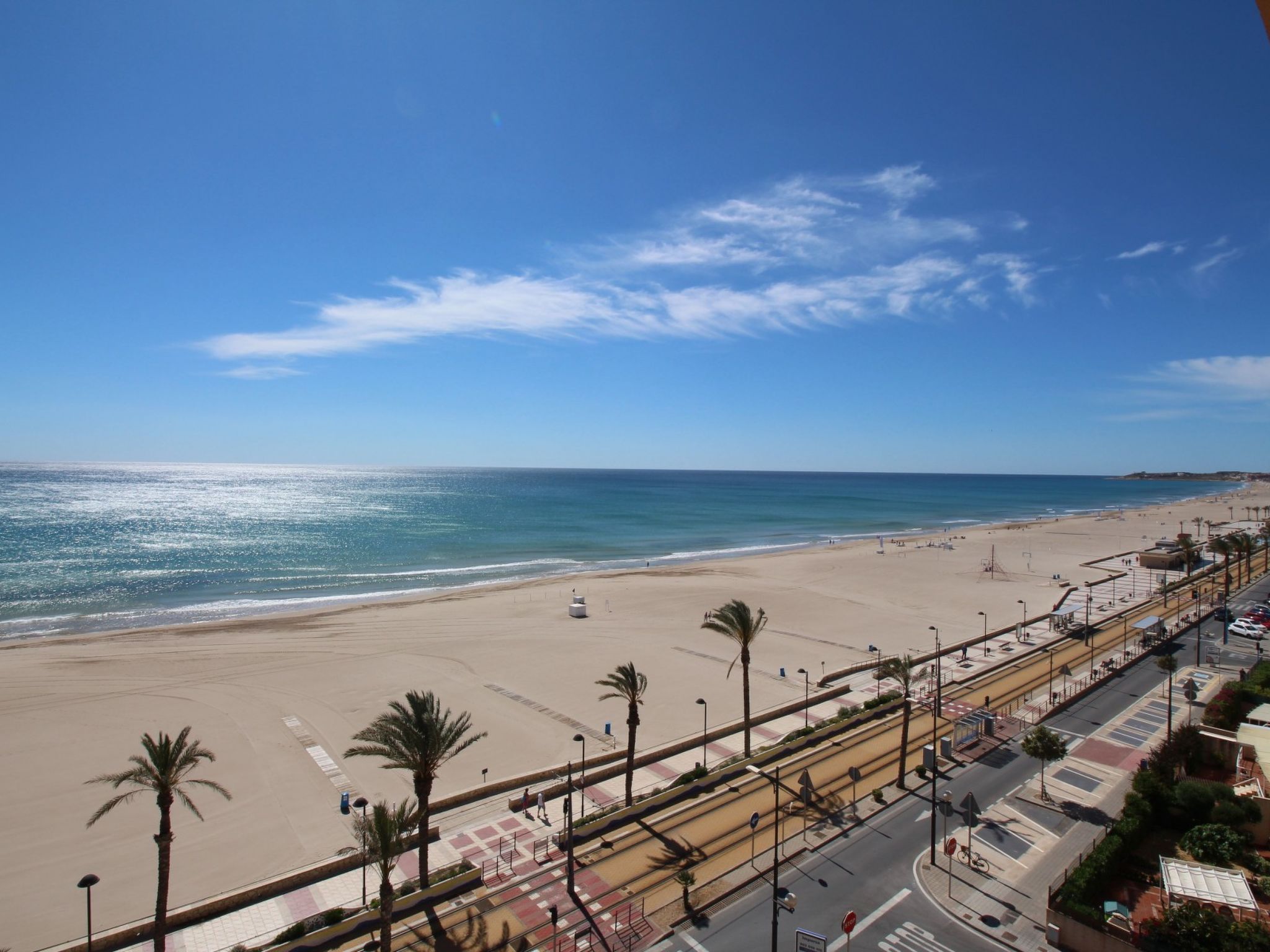 Une plage bordée d’une route parallèle au rivage, des palmiers et un ciel dégagé.