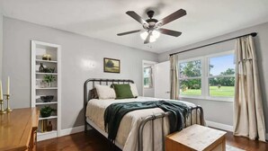 Een slaapkamer met een bed, een nachtkastje, een plafondventilator, een raam met gordijnen en een inbouwkast met decoratieve objecten.