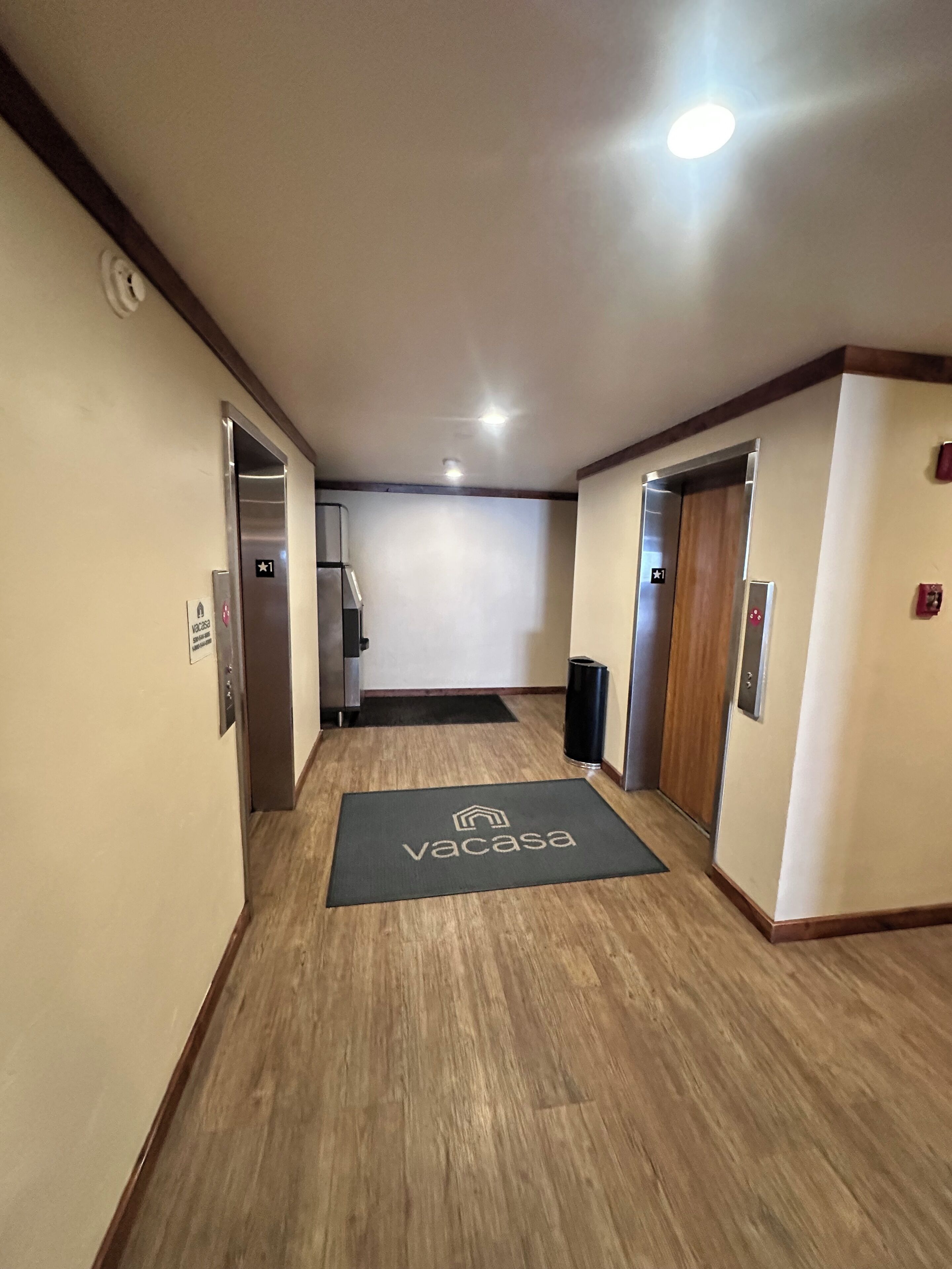 Un pasillo con piso de madera, dos ascensores, un felpudo negro con el logotipo de 'Vacasa' y un intercomunicador en la pared.