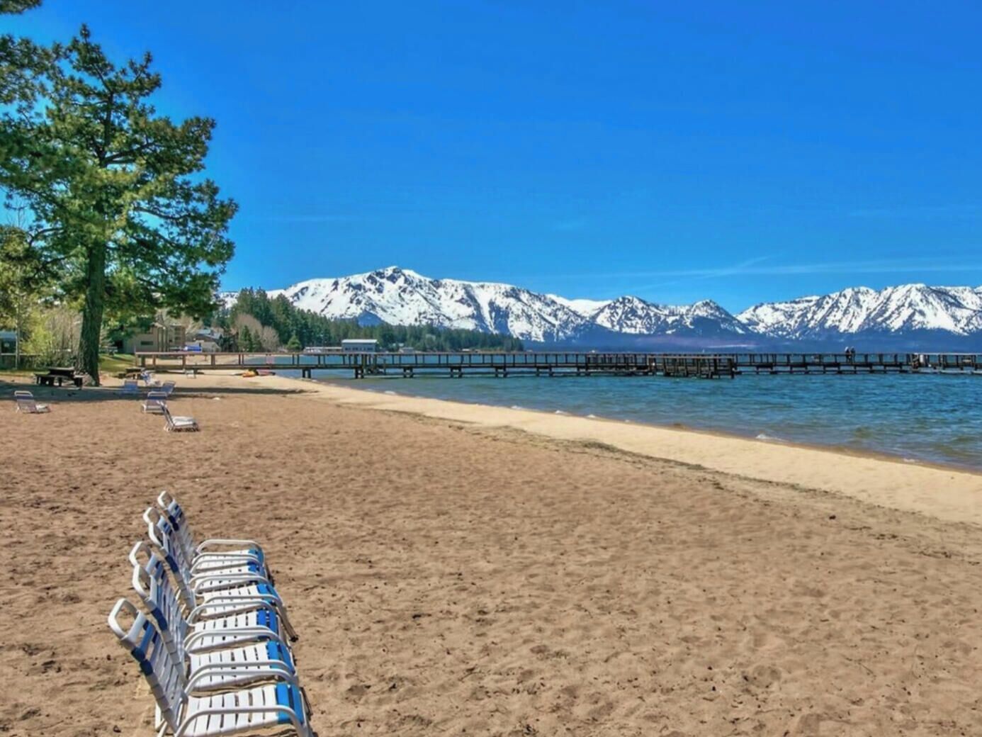 Ein Strand mit einer Reihe von Liegestühlen, einer hölzernen Promenade, die sich ins Wasser erstreckt, und schneebedeckten Bergen im Hintergrund.