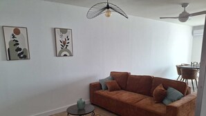 Een woonkamer met een bruine bank, twee ronde tafels met geometrische poten, een plafondventilator en twee aan de muur hangende kunstwerken.