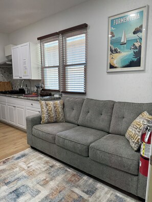 Sala de estar con un sofá gris, cojines estampados, un área de cocina con armarios blancos, una ventana con persianas y un cuadro en la pared.
