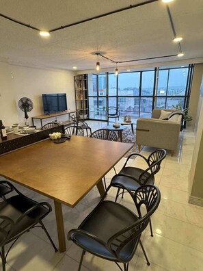 A modern kitchen with a large island, dining chairs, a TV, and a view of the city through large windows.