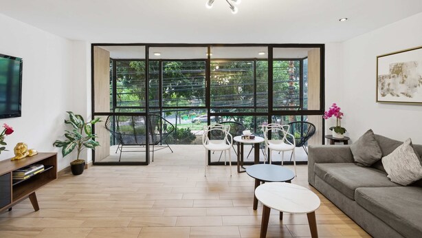 Uma sala de estar moderna com sofá, mesa de centro e cadeiras, janelas amplas com vista para o verde e um lustre.