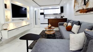 Ein modernes Wohnzimmer mit Flachbildfernseher, grauem Sofa, Couchtisch und einer Küche im Hintergrund.