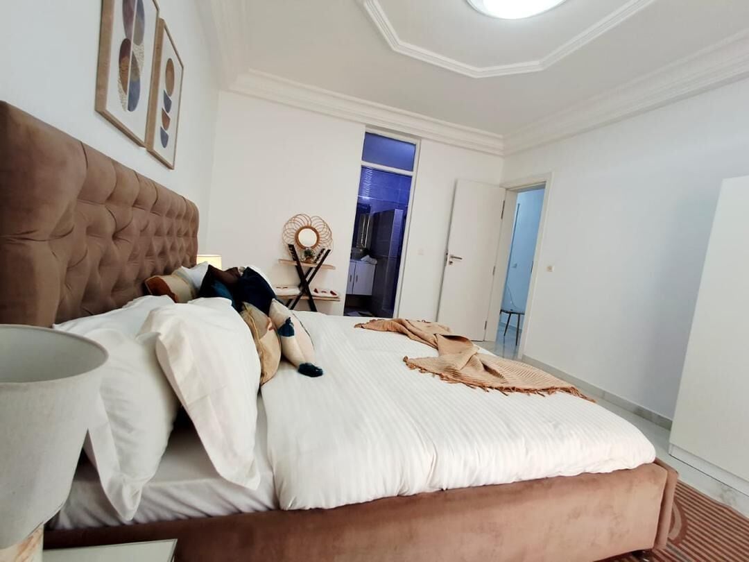 Ett sovrum med en stoppad sänggavel, en säng med vita sängkläder, ett sängbord med en lampa och en dekorativ väggdekoration.