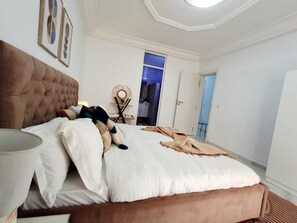 A bedroom with a tufted headboard, a bed with white bedding, a bedside table with a lamp, and a decorative wall piece. - Espace Virage type F4 (Dakar)