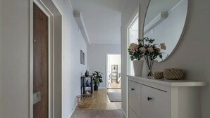 Ein Flur mit einer Holztür, einer weißen Wand, einem runden Spiegel, einem weißen Schrank, einem Korbwäschekorb und einer Topfpflanze.