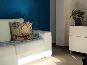 Ein modernes Wohnzimmer mit einem weißen Sofa, einem dekorativen Kissen, einer kleinen Pflanze auf einem weißen Schrank und einer blauen Wand.