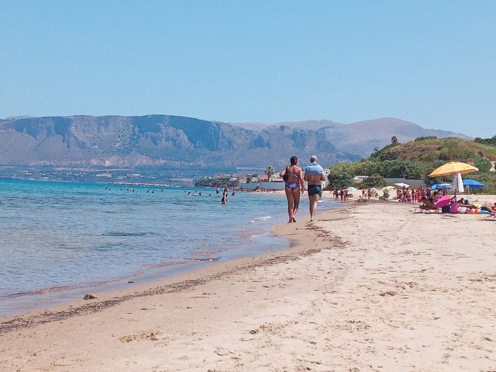 A beach with people walking, umbrellas, and a mountainous backdrop.
