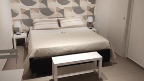 Un dormitorio con cama, mesitas de noche, un banco, un papel tapiz estampado y una puerta.
