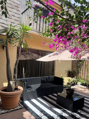 Eine Terrasse mit einer schwarzen Rattan-Couch, einer großen Topfpflanze und einem hängenden Pflanzgefäß mit rosa Blüten.