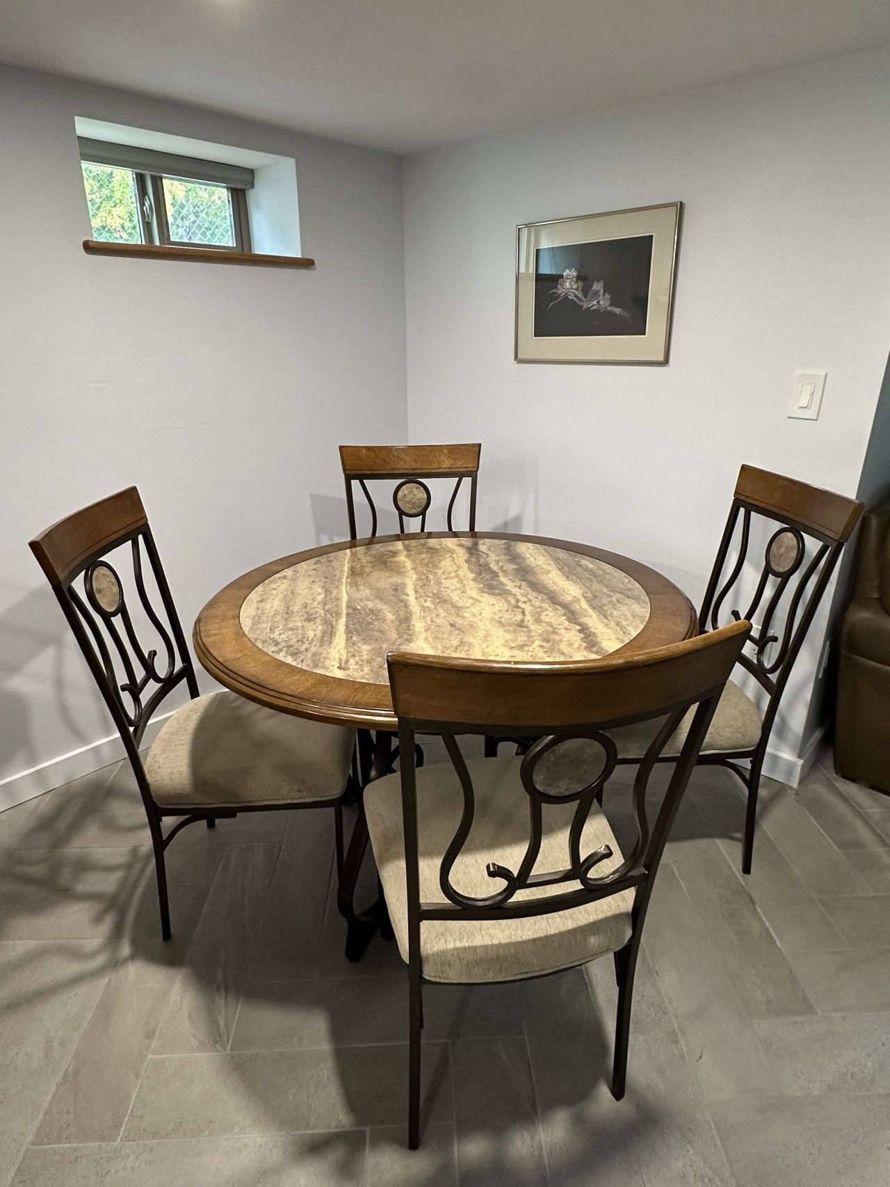 A dining area with a round table and four chairs, a framed picture on the wall, and a window with blinds.