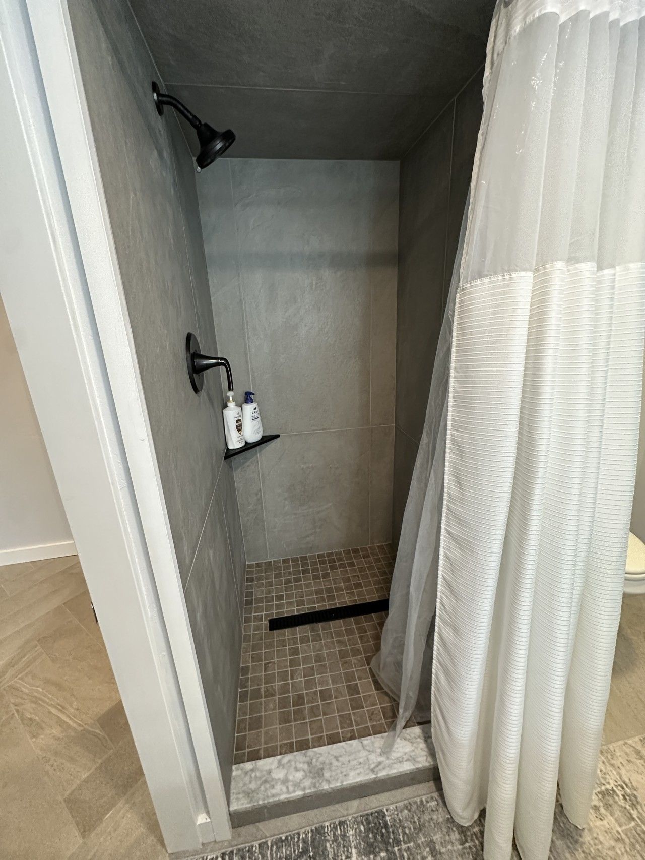 A shower area with a tiled floor, a showerhead, and a shelf with toiletries.