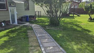 Un chemin mène à une maison avec un petit arbre et un barbecue dans la cour.