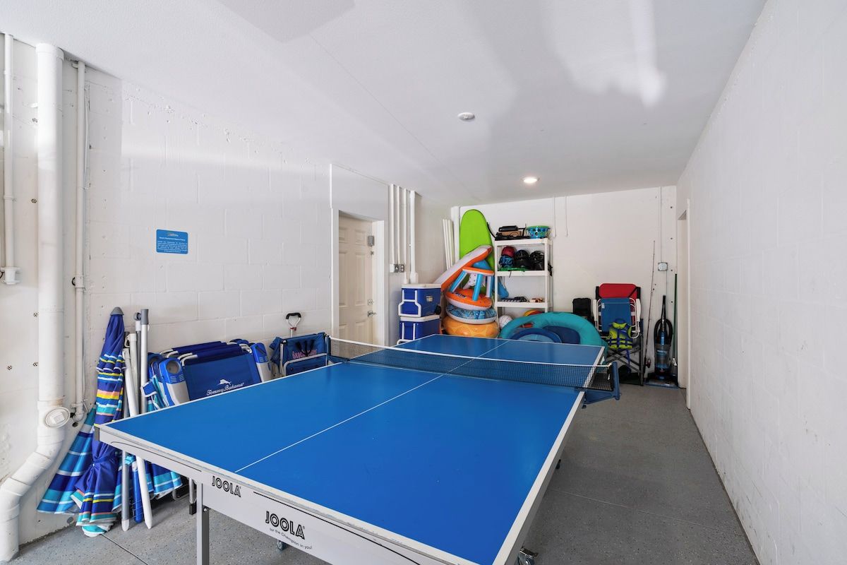 Una mesa de ping pong en una habitación con paredes blancas, una puerta y varios equipos deportivos.