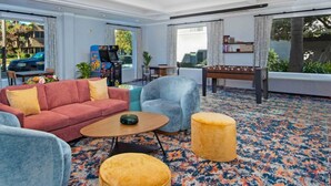 A hotel room with a red sofa, two armchairs, a small round table, a yellow ottoman, arcade games, a dining table, and a large window with a view.