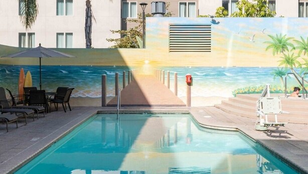A swimming pool with lounge chairs and umbrellas, a lifeguard stand, and a mural of a beach scene.