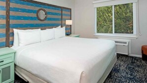 A hotel room with a large bed, a round mirror on the headboard, a window with blinds, and a small table with a lamp.