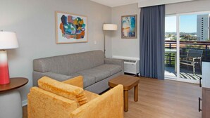 A hotel room with a grey sofa, wooden side table, a small wooden table, a TV, and a balcony with a view.