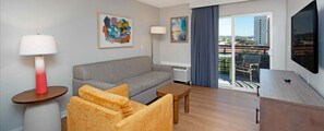Hotellihuone, jossa on harmaa sohva, puinen sivupöytä, pieni puinen pöytä, televisio ja parveke, jolta on näkymä.