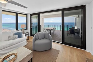 A modern living room with a view of the ocean, a balcony with a table and chairs, and a grey armchair. - 17th-Floor Gulf View Condo | Heated Pool, Hot Tub (Panama City Beach)