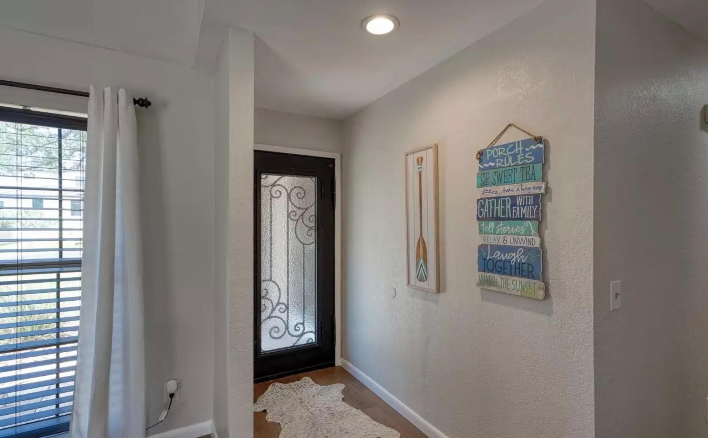 A hallway with a white door, a window with blinds, and a wall with a decorative sign.