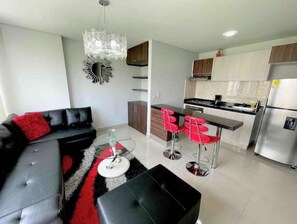 Ein modernes Wohnzimmer mit einer schwarzen Couch, roten Kissen, einem Glastisch und einem roten Teppich. Angrenzend befindet sich eine Küche mit Bar, Edelstahlgeräten und Holzausbau.