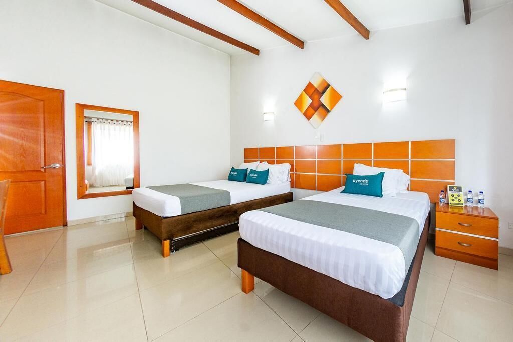A hotel room with two beds, a wooden door, a mirror, a wall-mounted light, and a small table with a phone and water bottles.
