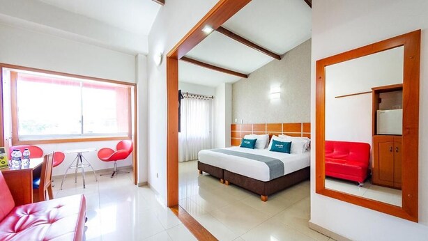 A hotel room with a bed, a red sofa, a dining area with chairs, and a window.